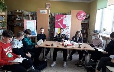 Dziewięcioro uczni&oacute;w siedzi w p&oacute;łokręgu przy stole w bibliotece i czyta książki
