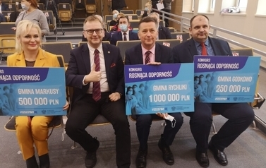 Cztery osoby siedzące na krzesłach, z kt&oacute;rych trzy prezentują otrzymane nagrody