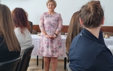 Kobieta stojąca pośr&oacute;d grupy innych siedzących os&oacute;b